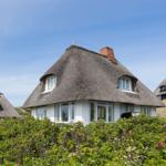 Dorfhaus mit Strohdach und weißen Wänden. Blumenbeet im Vordergrund.