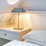Schlafzimmer mit Nachttisch, Lampe und Bett. Wandverkleidung und Steckdose sichtbar.