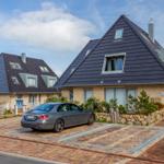 Modernes Ferienhaus mit dunklem Dach und Steinmauer. Ein Auto parkt auf dem gepflasterten Hof.
