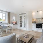 Living kitchen with dining area, sofa, and TV. White furniture, large windows.