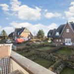 Balcony view of brick houses with gardens and stone walls under blue sky.