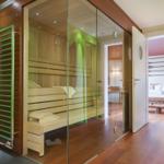 Sauna mit Glaswand, Holzbänken und grüner Wand. Blick in Schlafzimmer mit Bett.