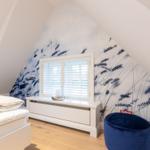 Schlafzimmer mit Dachschrägen, Fenster mit Jalousie und blauem Sitzkissen.