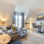 Living and dining area with sofa, chair, and table. Wooden floor and large windows.