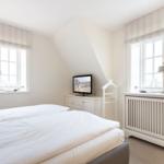 Schlafzimmer mit Doppelbett, zwei Fenstern und Fernseher auf Schrank.