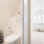 Badezimmer mit Toilette und kleinem Regal. Blick in Schlafzimmer mit Fenster und Stuhl.