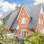 Rotes Backsteingebäude mit dunklem Satteldach und weißen Fenstern. Gartenbereich mit Sitzgelegenheit.
