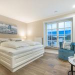 Schlafzimmer mit Blick aufs Meer, weißes Bett, blauer Sessel und Fenster mit Meerblick.