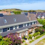 Haus mit dunkelblauem Dach und roten Ziegelsteinen, umgeben von grünen Bäumen und lila Blumen.