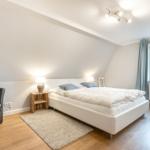 Bedroom with bed, armchair, and window. Wooden floor and white walls.