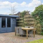 Garden house with terrace, table and chairs. Wooden fence with ivy. Paved path and grass area.