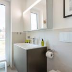 Badezimmer mit Waschbecken, Spiegel und Fenster. Wandverkleidung und Toilettenpapierhalter sichtbar.
