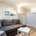 Wohnzimmer mit Sofa, Beistelltisch und Wandbildern. Durchgang zur Küche sichtbar.