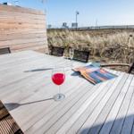 Holztisch mit zwei Gläsern und Zeitschrift auf Terrasse mit Strandhintergrund.
