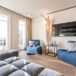 Wohnzimmer mit blauen Sitzmöbeln, Fernseher und großem Fenster zur Terrasse.