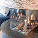 Ein moderner Kamin mit brennenden Holzscheiten und einem runden Tisch. Ein blauer Sessel ist im Hintergrund sichtbar.