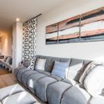 Wohnzimmer mit grauem Sofa, Kissen und Wandbildern. Blick in die Küche mit Esstisch.
