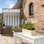 Terrasse mit Strandkorb, Blumen und rotem Backsteinhaus.