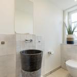 Badezimmer mit Steinwaschbecken, Spiegel und Toilette. Fenster mit Pflanze und Handtuch.