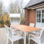 Terrasse mit Tisch, Stühlen und Strandkorb. Hintergrund: Haus mit Glasfront und Holzzaun.