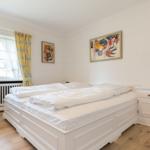 Schlafzimmer mit weißem Bett, gelben Vorhängen und abstraktem Bild an der Wand.