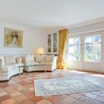 Wohnzimmer mit cremefarbenen Sofas, Glas-Tisch und Terrakottboden. Große Fenster mit gelben Vorhängen.