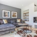 Ein Wohnzimmer mit grauem Sofa, Kamin und runden Tischen auf einem Fellteppich.