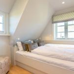 Schlafzimmer mit Dachschrägen, Bett, Fenstern und Sitzhocker.