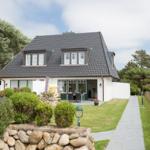 Haus mit dunklem Dach, weißer Fassade und Gartenweg. Terrasse mit Tisch und Stühlen.