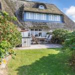 House with thatched roof, terrace with table and chairs, surrounded by grass and shrubs.