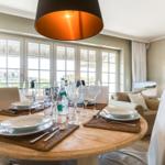 Round table with placemats and glasses. Windows with view. Living area in background.