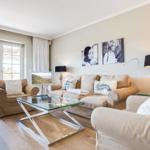 Living room with sofa, armchairs, and glass coffee table. Two pictures hang on the wall.