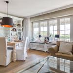 Dining area with table and chairs, living area with sofa and glass table, large windows with view outside.