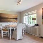 Dining room with table, chairs, and large windows. Wall art shows landscapes.