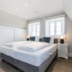 Schlafzimmer mit Doppelbett, zwei Fenstern und Nachttischen.