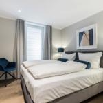 Schlafzimmer mit Doppelbett, blauer Sessel und Fenster mit Jalousie.