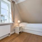 Schlafzimmer mit Dachschräge, weißem Bett und Holzboden. Fenster mit Vorhang und Heizkörper.
