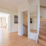House with wooden staircase, mirror, and view into living area with sofa and window.