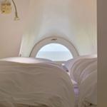 Zimmer mit Blick auf Meer durch runden Fenster, Bett mit weißen Bettwäsche, Wandlampe