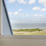 Durch das Fenster blickt man auf das Meer und grüne Dünen.