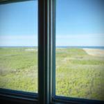Durch das Fenster blickt man auf eine grüne Dünenlandschaft und das Meer.