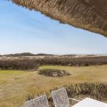 Terrace with thatched roof and white chairs overlooks dunes and beach.