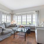 Wohnzimmer mit Sofa, Sessel und Glas-Tisch. Große Fenster mit Blick auf Grünfläche.