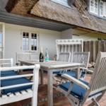 Terrasse mit Holztisch, Stühlen und blauen Kissen unter strohgedecktem Dach.