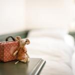 Ein Stoffmaus mit Koffer sitzt auf einem Tisch vor einem Bett.