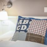 Ein Kissen mit Patchwork-Muster liegt auf einem Bett. Eine Wandlampe ist im Hintergrund sichtbar.
