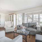 Wohnzimmer mit grauem Sofa, weißem Sessel und Glas-Tisch. Große Fenster und Terrasse.