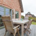 Terrasse mit Holztisch und Stühlen vor einem Backsteinhaus mit grünen Fenstern.