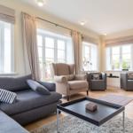 Wohnzimmer mit Sofa, Sesseln und Fenstern mit Vorhängen.