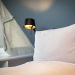 Bettkopf mit Kissen, Wandlampe und Fenster mit Vorhang.
